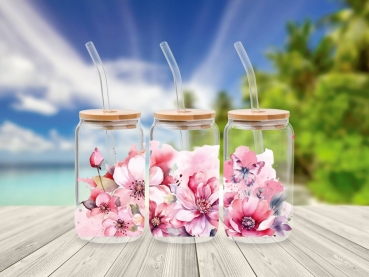 Drei 16 oz Glasbecher mit Bambusdeckel und Glastrinkhalm, verziert mit rosa und pinkfarbenen Blumenmotiven und einem Schmetterling. Die Becher stehen auf einem Holztisch vor einem tropischen Strandhintergrund, was eine frische und sommerliche Atmosphäre v